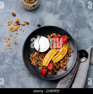 Oatmeal granola with yoghurt, fresh strawberries and banana, chia seeds, sunflower and honey in black ceramic plate on dark background. Top view. Stock Photo
