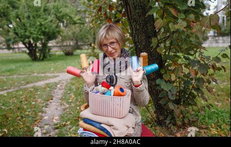 a seamstress woman is sitting on a chair on the street wearing coils with multicolored threads on her fingers. Stock Photo