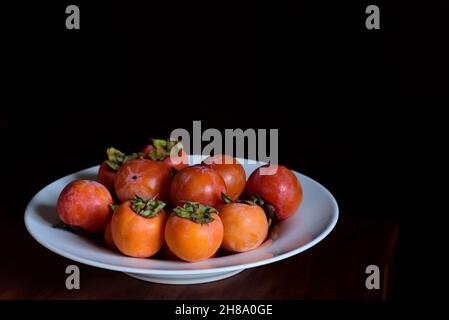 A plate of ripe persimmons stands on a wooden table against a dark background