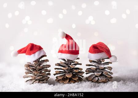 Three pine cones in a red Santa hats like a Christmas tree with bokeh lights in the snow on a white background, Christmas concept. Stock Photo
