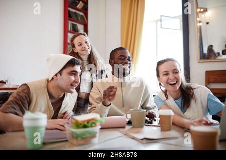 Group of friends laughing and looking something on laptop while sitting at the table in the room Stock Photo