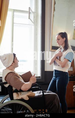 Young woman holding book and talking to man who using wheelchair, they doing homework together in the room Stock Photo