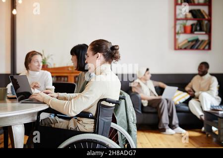 Student sitting in wheelchair and using laptop in his online study with his classmates at the table Stock Photo