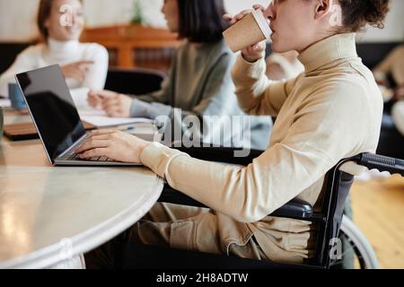 Student with disability using wheelchair sitting at the table and drinking coffee while using laptop Stock Photo