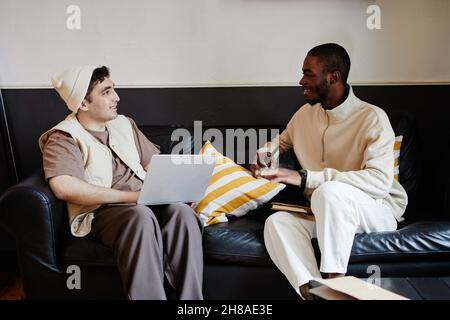 Two friends sitting on sofa with books and laptop and talking, they preparing for exams together Stock Photo