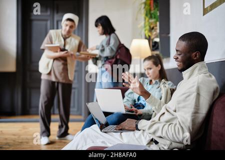 African happy student waving and smiling at camera while sitting on the sofa and talking online on laptop with other students in the bakground Stock Photo