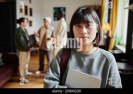 Portrait of Asian student with backpack and textbook looking at camera standing in the classroom with students in the background Stock Photo