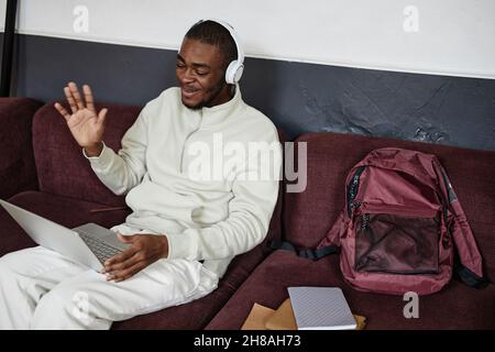 African student in headphones waving to the camera while studying online on laptop sitting on sofa Stock Photo