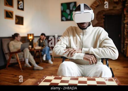African young man in vr glasses sitting in front of the chessboard and playing chess virtually Stock Photo
