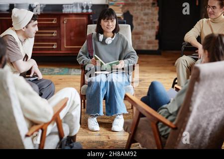 Young woman sitting in armchair and reading a book for group of students during classes Stock Photo