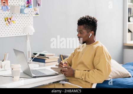 Serious young student of African ethnicity making notes while listening to teacher on laptop display at lesson Stock Photo