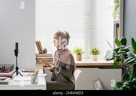 Young confident teacher in hijab looking in smartphone camera while explaining something to students during livestream Stock Photo