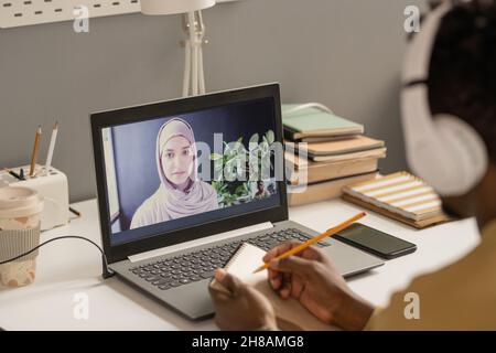 Young teacher in hijab on laptop screen looking at African guy with notepad and pencil making notes during online lesson Stock Photo