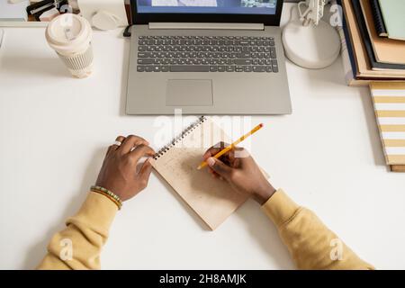 Hands of young African man writing down algebraic formula on page of notepad while sitting by desk in front of laptop Stock Photo