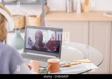 Young Muslim female with financial document on laptop screen explaining data to student during online lesson Stock Photo