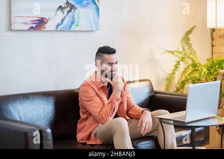 Happy young man sitting on sofa in front of laptop and communicating with teacher during online lesson Stock Photo