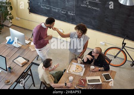 Group of intercultural young colleagues eating pizza and discussing plans for evening while having lunch break by table in office Stock Photo
