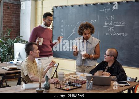 Group of young contemporary colleagues having pizza and drinks during discussion of working points at lunch break Stock Photo