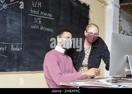 Two young office workers in protective masks looking at computer screen during discussion of financial data at meeting Stock Photo