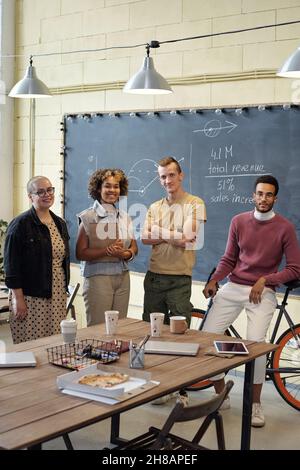 Happy young economists standing by table with drinks, pizza and gadgets in large openspace office and looking at camera Stock Photo