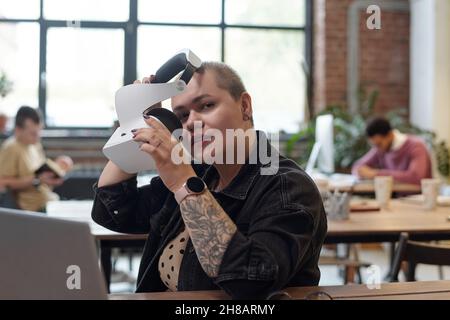 Young businesswoman in casualwear putting on vr headset while sitting by workplace against two male co-workers Stock Photo