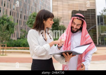 Arab man client and caucasian businesswoman discussing contract details at outdoor meeting, happy saudi businessman singing document on clipboard while standing on city street with female secretary  Stock Photo