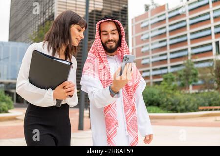 Two diverse business colleagues arab man and caucasian woman looking at smartphone screen with smile while standing on city street, using phone to search information together in internet Stock Photo