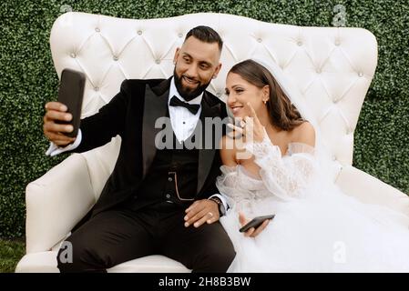 Smiling bride and groom sitting on sofa, taking selfie Stock Photo