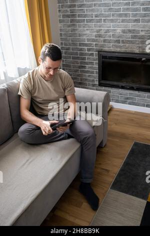 A young man is sitting on a sofa and playing on a tablet. Guy typing something Stock Photo