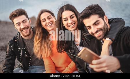 Group of gen z young people enjoying weekend sitting seaside on a bench having fun watching social network content on their smartphone - people using Stock Photo