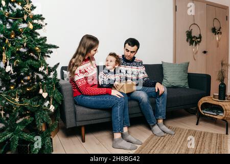 Happy family sitting on sofa near Christmas tree at home. Christmas sweaters. Stock Photo