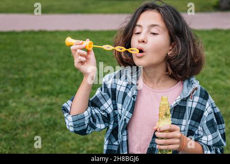 Attractive schoolgirl of Caucasian ethnicity blowing soap bubbles. Portrait of a cute beautiful brunette girl having fun in a summer park. Positive em Stock Photo