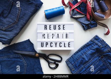 Reuse, repair, upcycle text on light board on sewing machines background. Stack of old jeans, Denim clothes, scissors, thread and sewing tools in Stock Photo