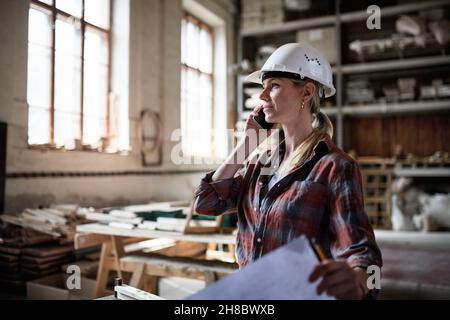 Female engineer holding blueprints and making phone call indoors in carpentry workshop. Stock Photo