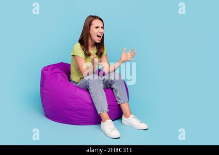 Full size photo of mad brunette millennial lady sit on bag watch tv wear t-shirt jeans shoes isolated on blue color background Stock Photo