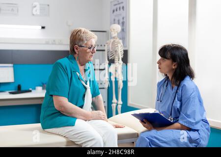Medical assistant consulting aged woman with physical injury in office. Nurse doing examination for orthopedic diagnosis and recovery, talking about pain relief and physiotherapy Stock Photo