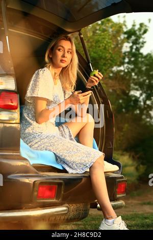 Blonde in a blue dress has a rest sitting in the open trunk of a car and a bite apple. A trip alone near the house in the countryside. Stock Photo