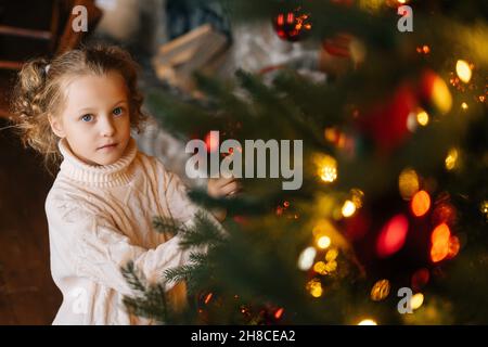 High-angle view of pretty little blonde curly child girl decorating Christmas tree with festive balls and toys in cozy living room looking at camera. Stock Photo