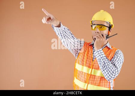 Portrait of contractor in hardhat, protective glasses and orange vest talking on walkie-talkie and pointing with finger Stock Photo