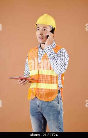 Serious young construction engineer in hardhat holding tablet computer and talking on phone with coworker Stock Photo