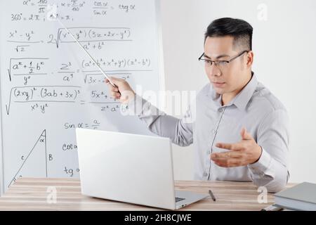 Serious high school teacher pointing at whiteboard with formulas when explaining new topic sduring online lesson Stock Photo