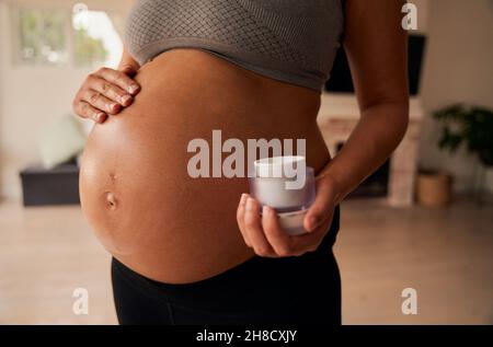 Caucasian female standing in living room rubbing moisturiser onto pregnant belly  Stock Photo