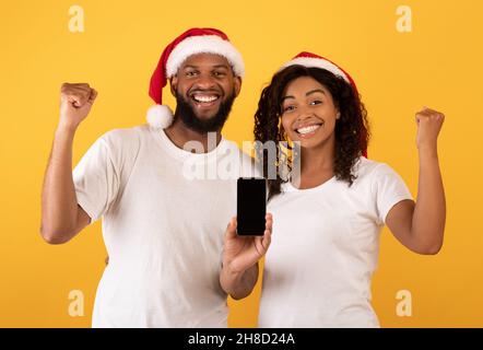 Yes. Excited black couple holding smartphones with empty screen shaking clenched fists, yellow background, mockup Stock Photo