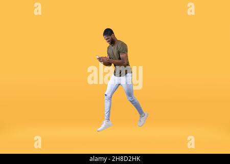 Young african american man jumping and using smartphone over yellow background, full-length portrait Stock Photo