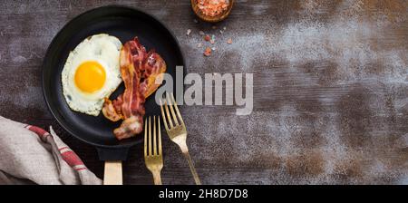 Traditional English breakfast with fried eggs and bacon in cast iron pan on dark concrete background. Top view. Stock Photo