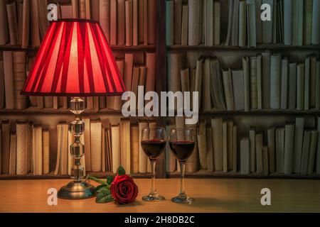 Table lamp with red shade, two glasses filled with red wine and a single red roses in front of a bookself Stock Photo