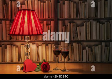 Table lamp with red shade, two glasses filled with red wine, single red roses and red hearts in front of a bookself Stock Photo