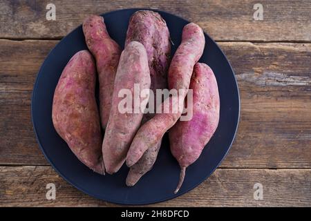 Sweet potato on black plate on wooden background. Raw potato tubers top view. Prepare on kitchen table. Sweet potatos for food with copy space. Stock Photo