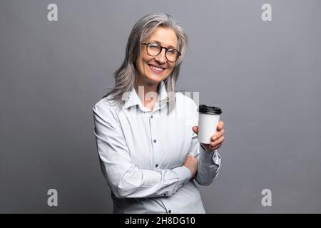 Morning coffee to go. Charming senior woman in formal wear standing with paper cup of hot drink isolated on gray, preparing to good working day, mature business lady takes a coffee break Stock Photo