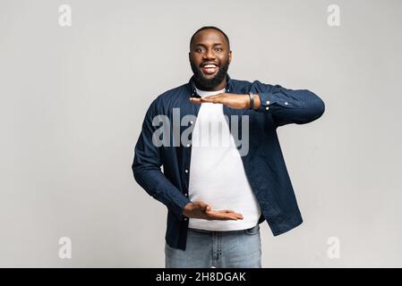 Portrait of smiling young handsome African American guy in blue shirt standing isolated over gray background and looking at the camera, showing measuring size with hands, copy space mockup image Stock Photo